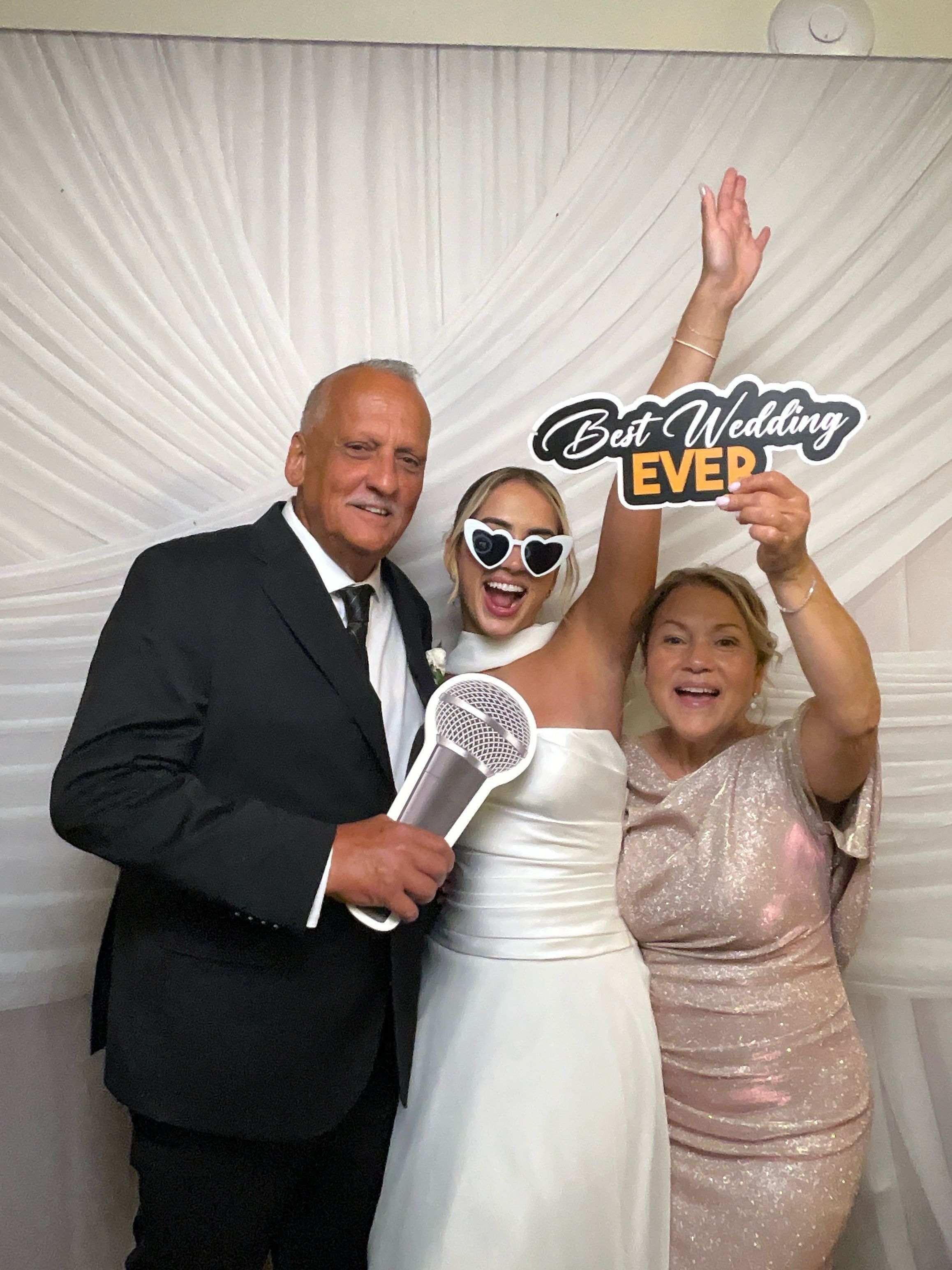 Bride in white dress with heart sunglasses poses with two guests holding Best Wedding Ever sign at wedding photo booth.