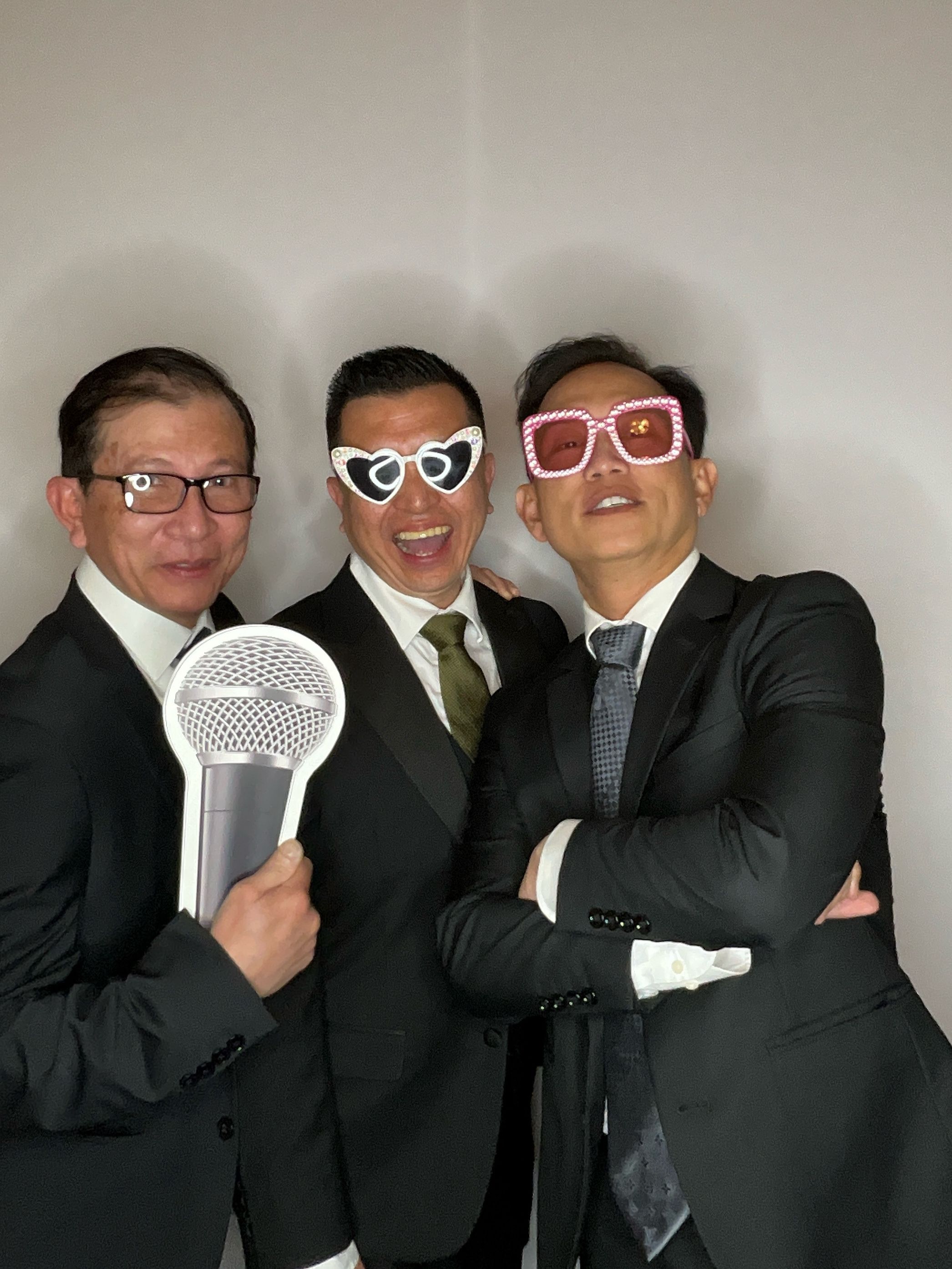 Three people in business suits pose playfully with sunglasses and a microphone prop in a photo booth sequence.