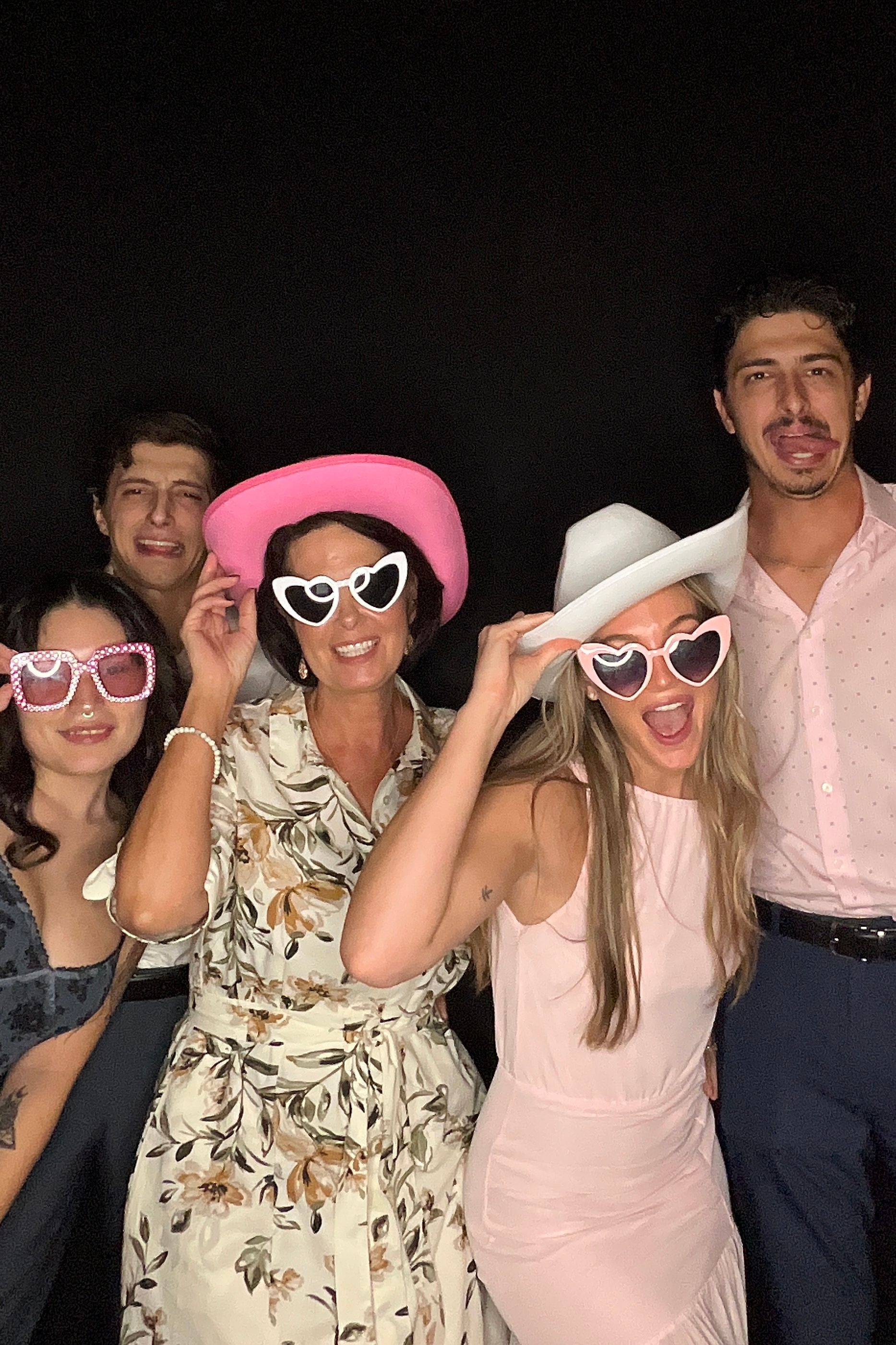 A group poses together at a nighttime event wearing sunglasses and casual attire including floral and pink outfits.