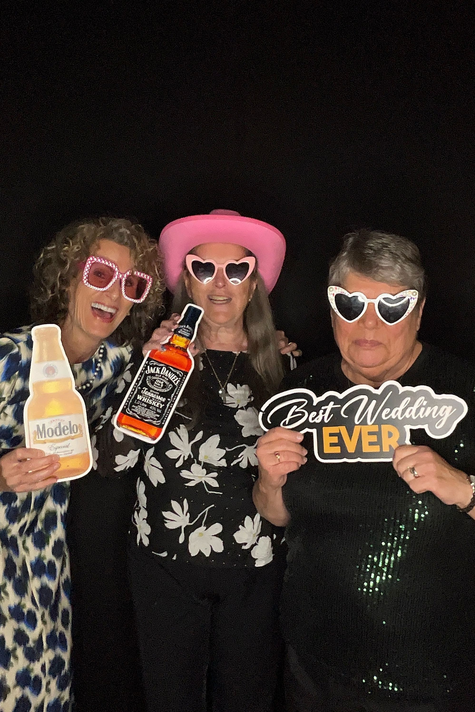 Three people wearing sunglasses pose with props including a Jack Daniels bottle and Best Wedding Ever sign at a photo booth.