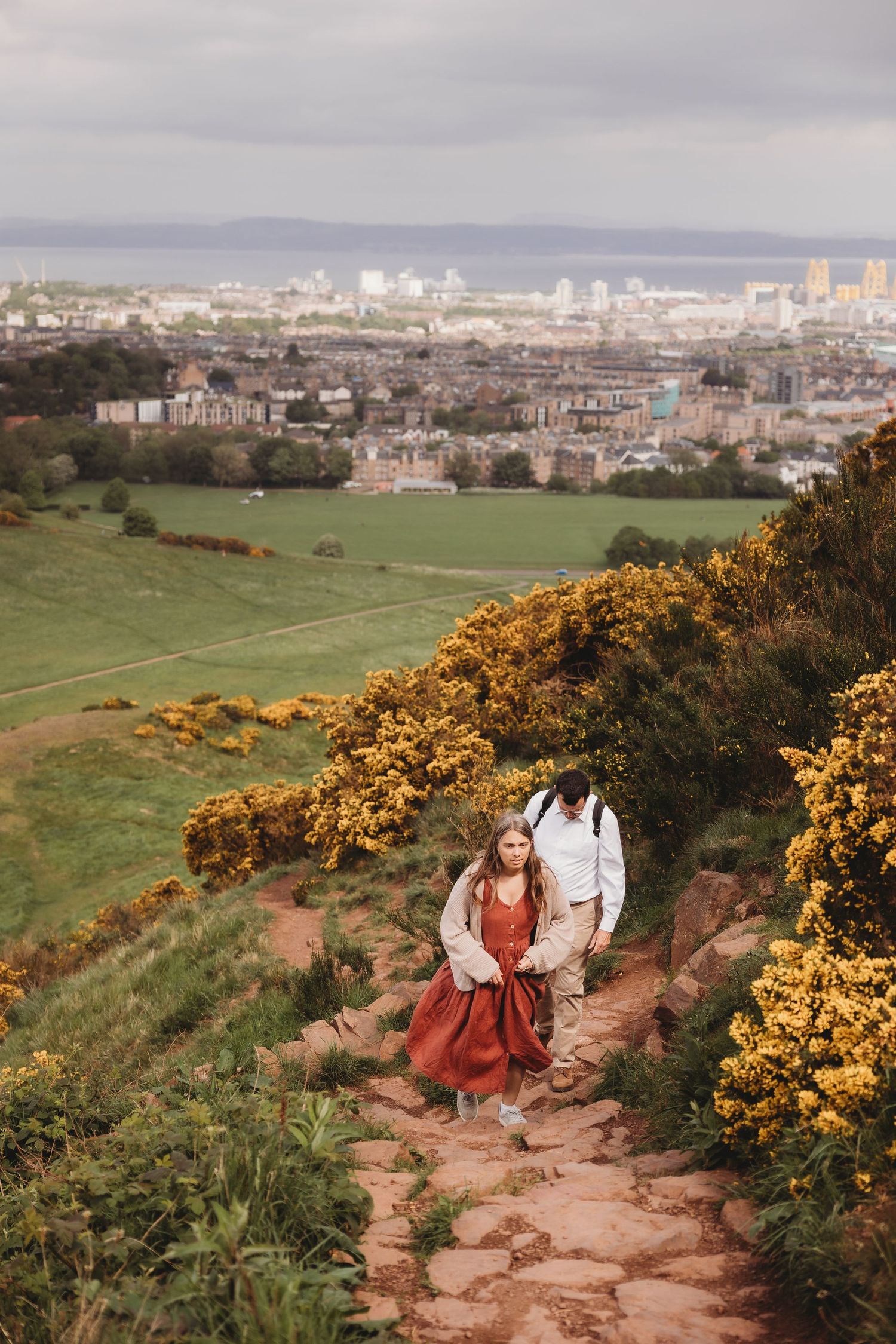Couple hiking a scenic trail at Arthur's Seat in Edinburgh, surrounded by yellow gorse bushes with city views behind.