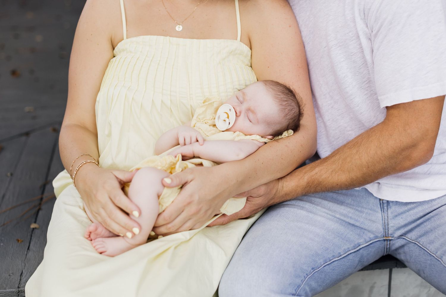 A mother wearing a pale yellow spaghetti dress holds her infant in a matching dress while father hugs her from the side while both sit.