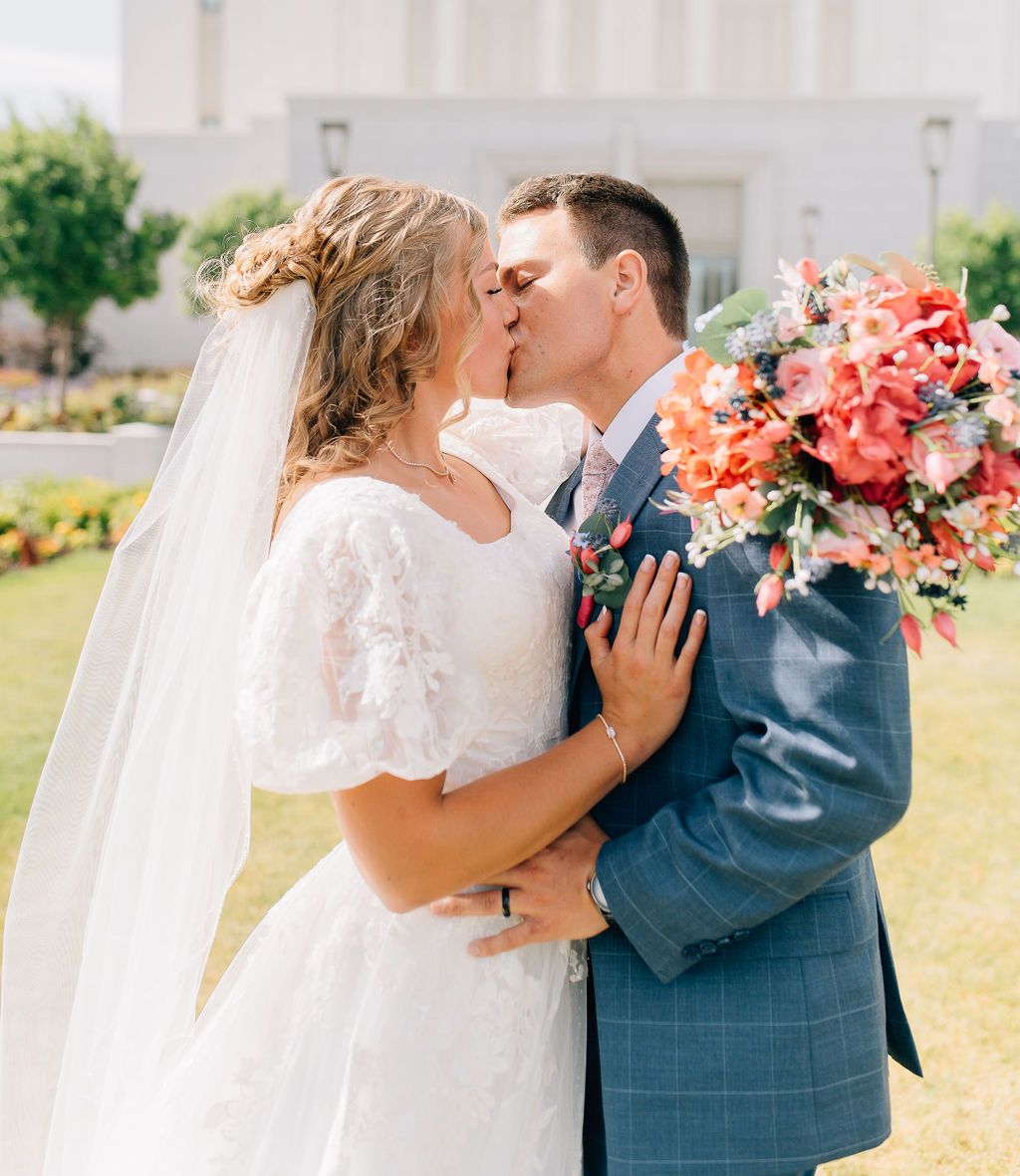 A couple shares a romantic kiss while holding a vibrant coral and pink floral bouquet in an outdoor garden setting.