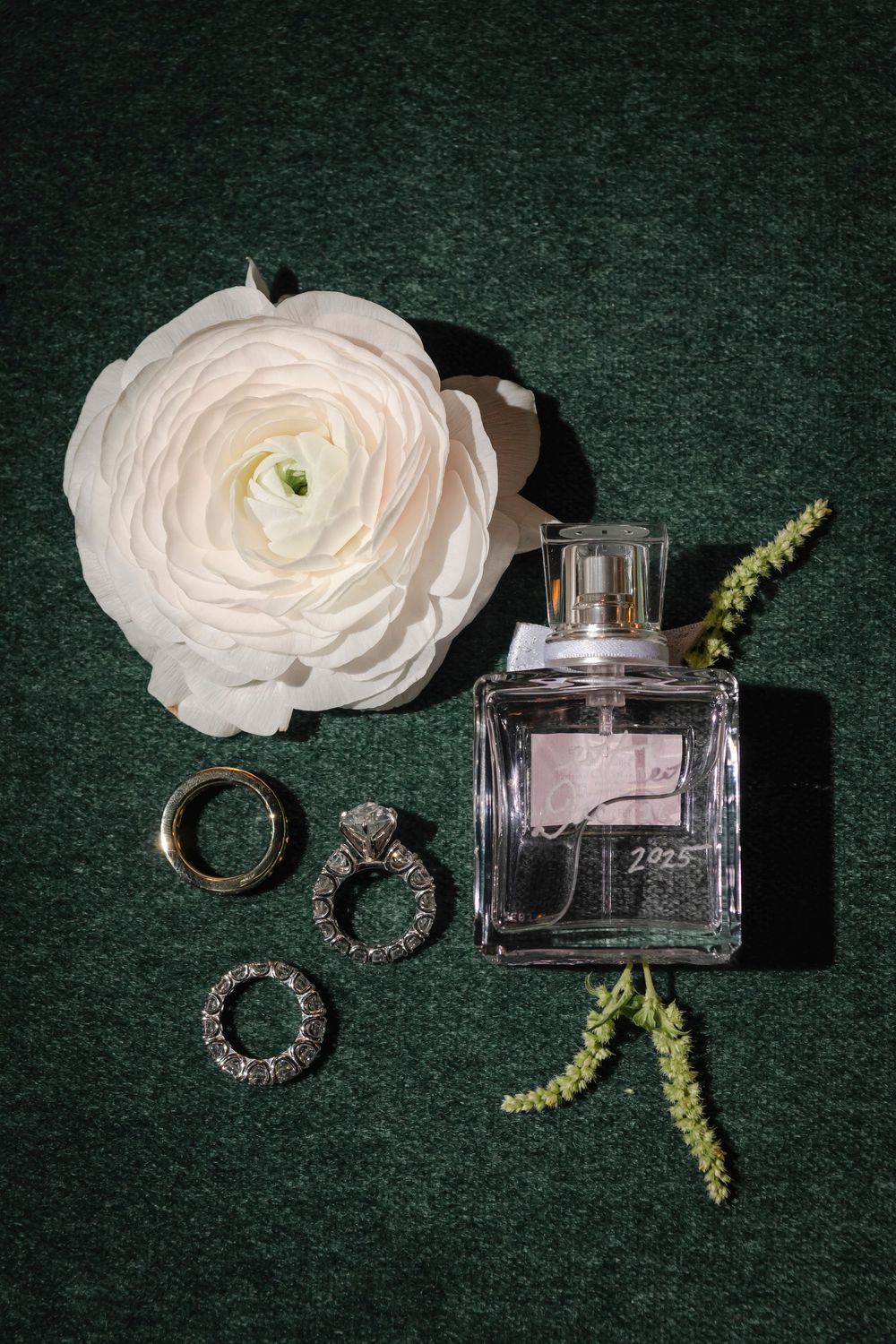 Elegant perfume bottle with silver rings and white ranunculus flower arranged on dark green velvet background.