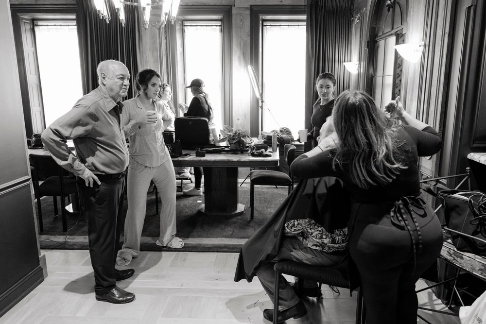 A black and white candid photo of a room with people getting hair and makeup done at different stations.