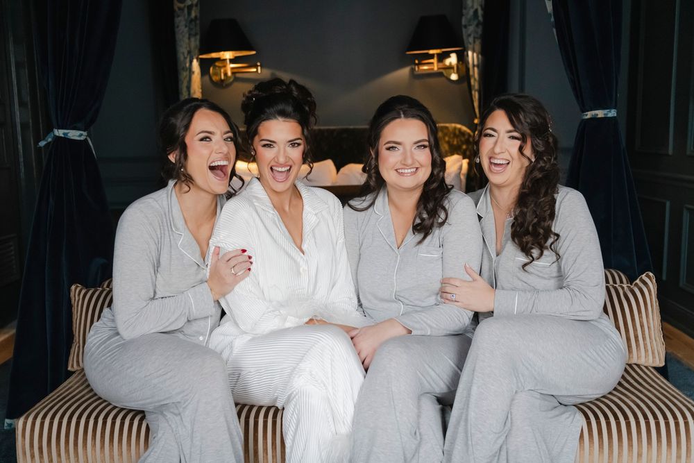 A bride and her 3 bridesmaids wearing light colored clothing laugh and smile together on a striped couch.