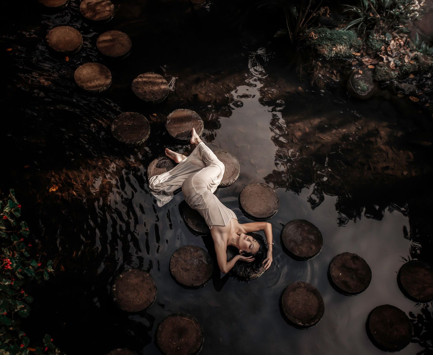Artistic dark photo of a figure lying in shallow water surrounded by lily pads.
