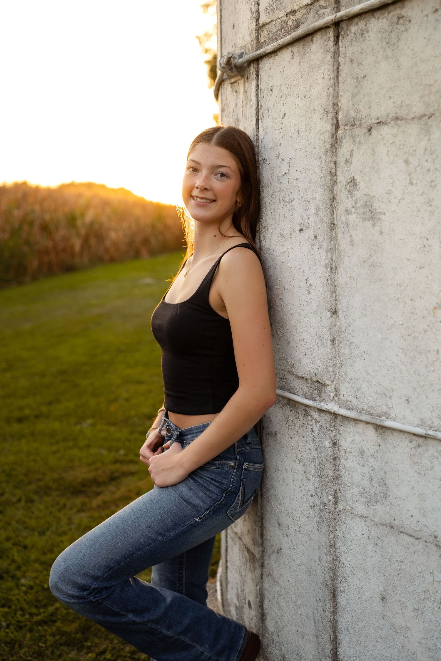 Person in black tank top and jeans poses against white wall in golden sunset light.