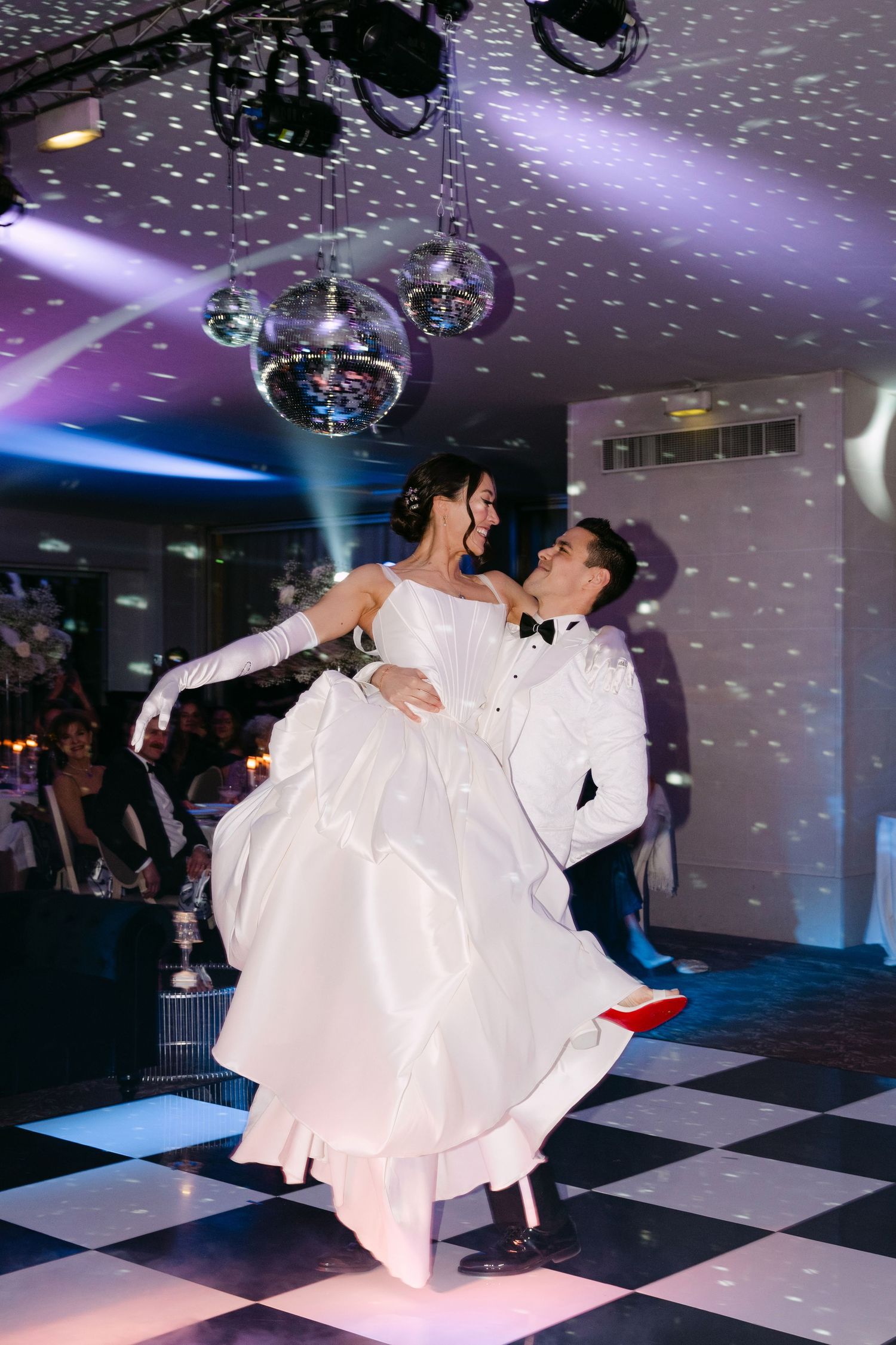 A couple performs an elegant dance dip on a black and white checkered dance floor under sparkling lights at a wedding reception.