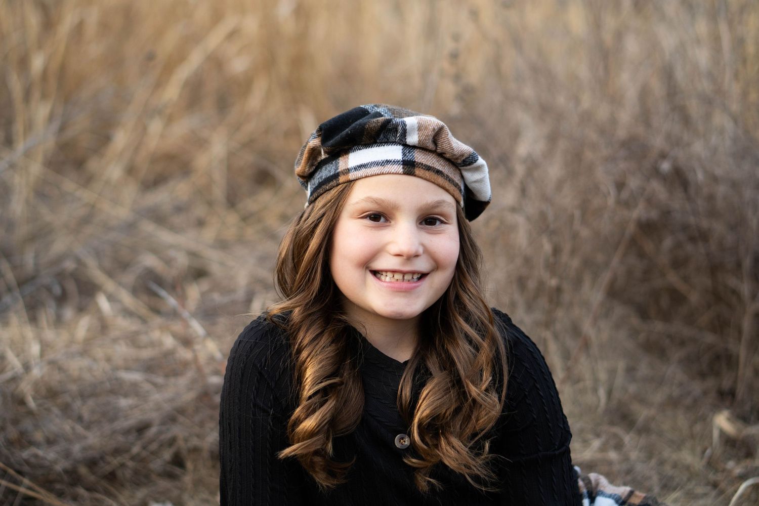 A smiling young girl with long brown hair wearing a plaid beret poses in a field of tall dry grass.