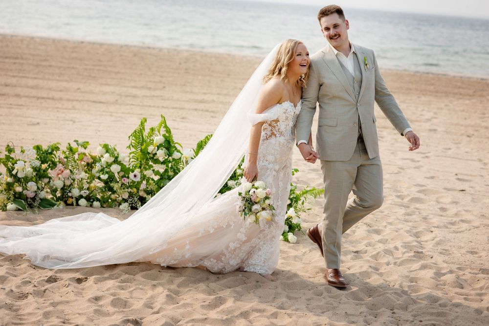 A couple walks together on a sandy beach during their Michigan wedding ceremony with floral decor and waterfront backdrop.