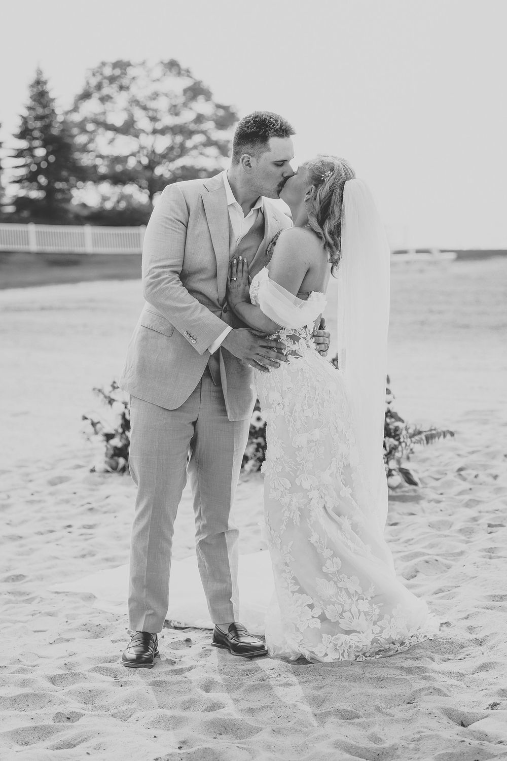 A romantic black and white beach wedding photo series showing a couple in formal attire embracing on the sand.