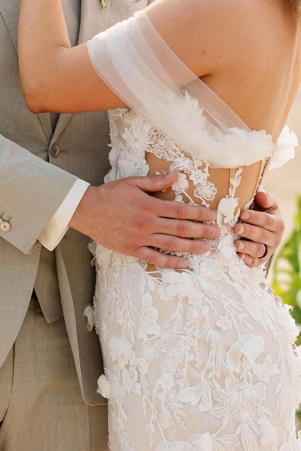 Close-up view of newlywed couple embracing, showing intricate lace details of white wedding dress and grey suit.