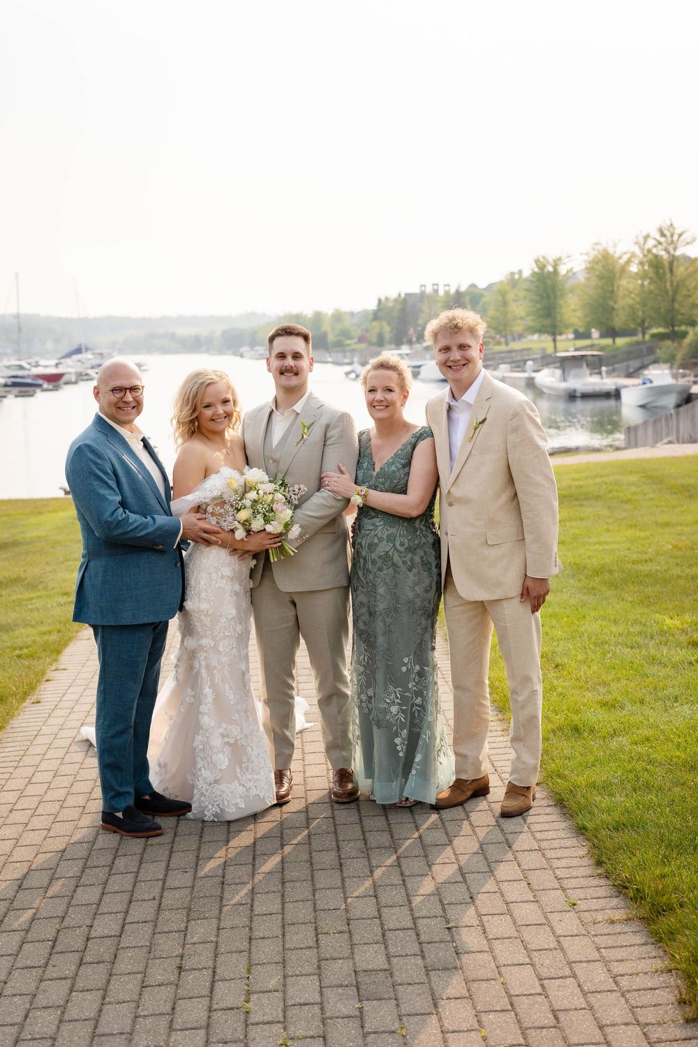 Wedding party poses together on stone patio with water and trees in background during outdoor celebration.