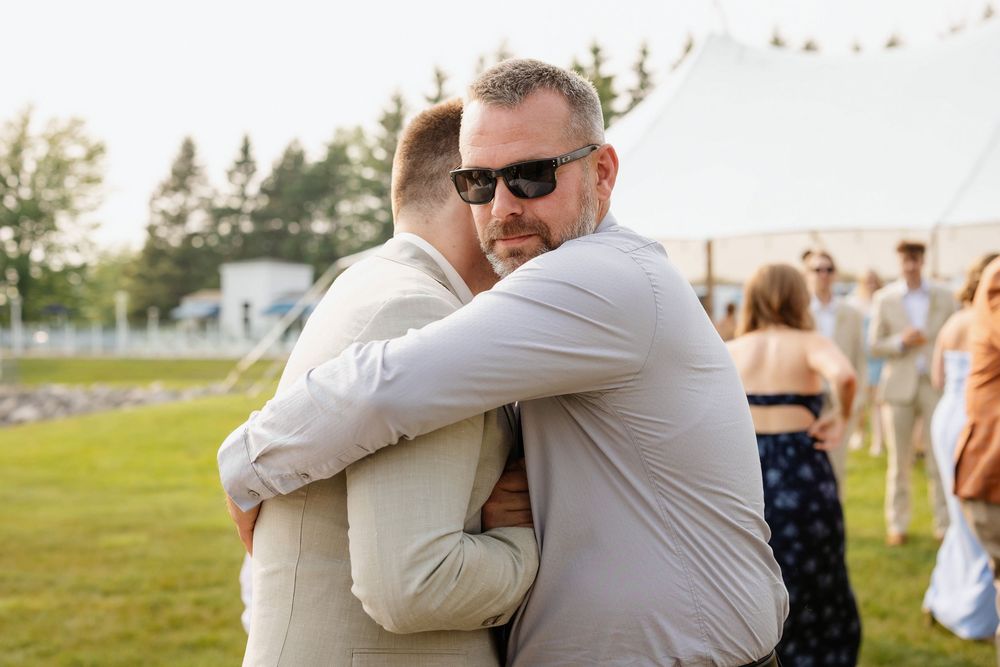 Two people share a warm embrace on a sunny lawn with a white tent and guests in the background.