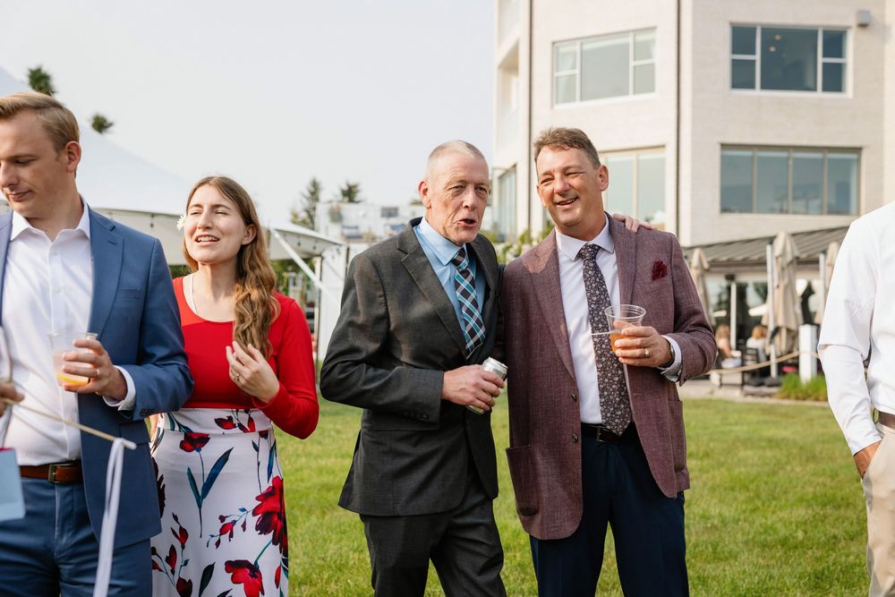 Wedding guests gather on a grassy lawn outside, laughing and enjoying drinks during an outdoor reception celebration.