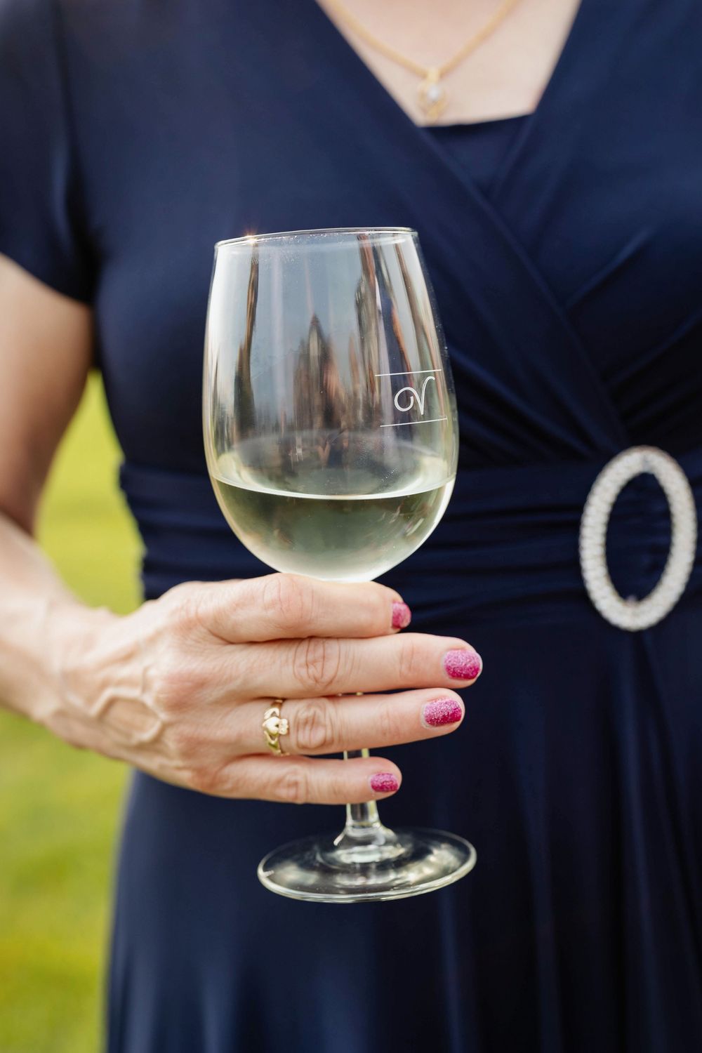 A hand wearing gold rings and pink nail polish holds a glass of white wine against a navy blue dress.