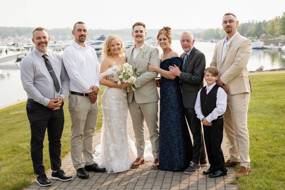 Wedding party in light beige and navy attire poses together on a grassy lawn by the water.