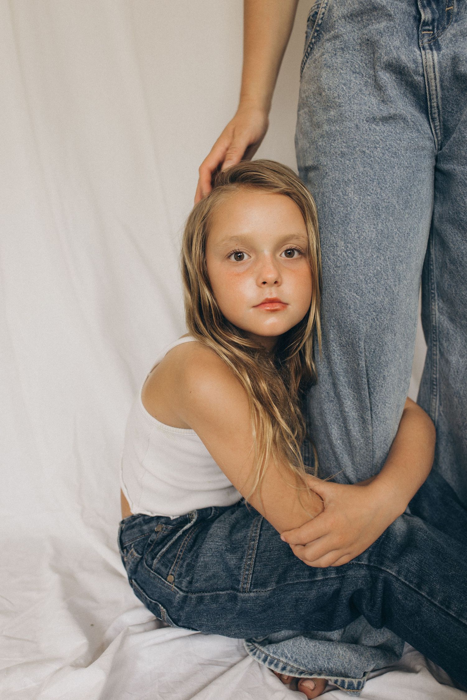 Young blonde girl in white tank top hugs adult's denim-clad legs while sitting on white fabric floor in New Zealand.
