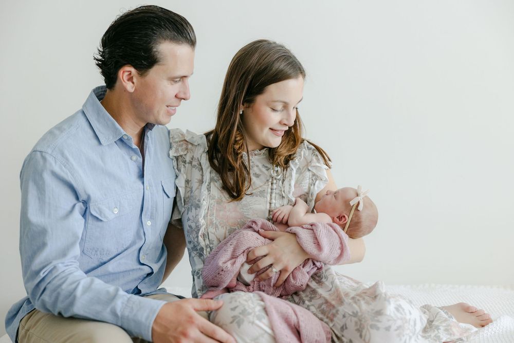 A family sits together on a couch, lovingly gazing at their newborn baby wrapped in pink blankets.