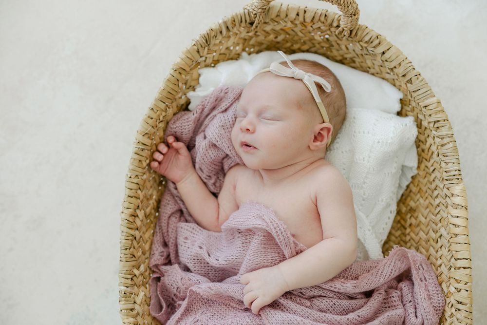 Sleeping newborn baby in pink dress rests peacefully in woven basket on white blanket.