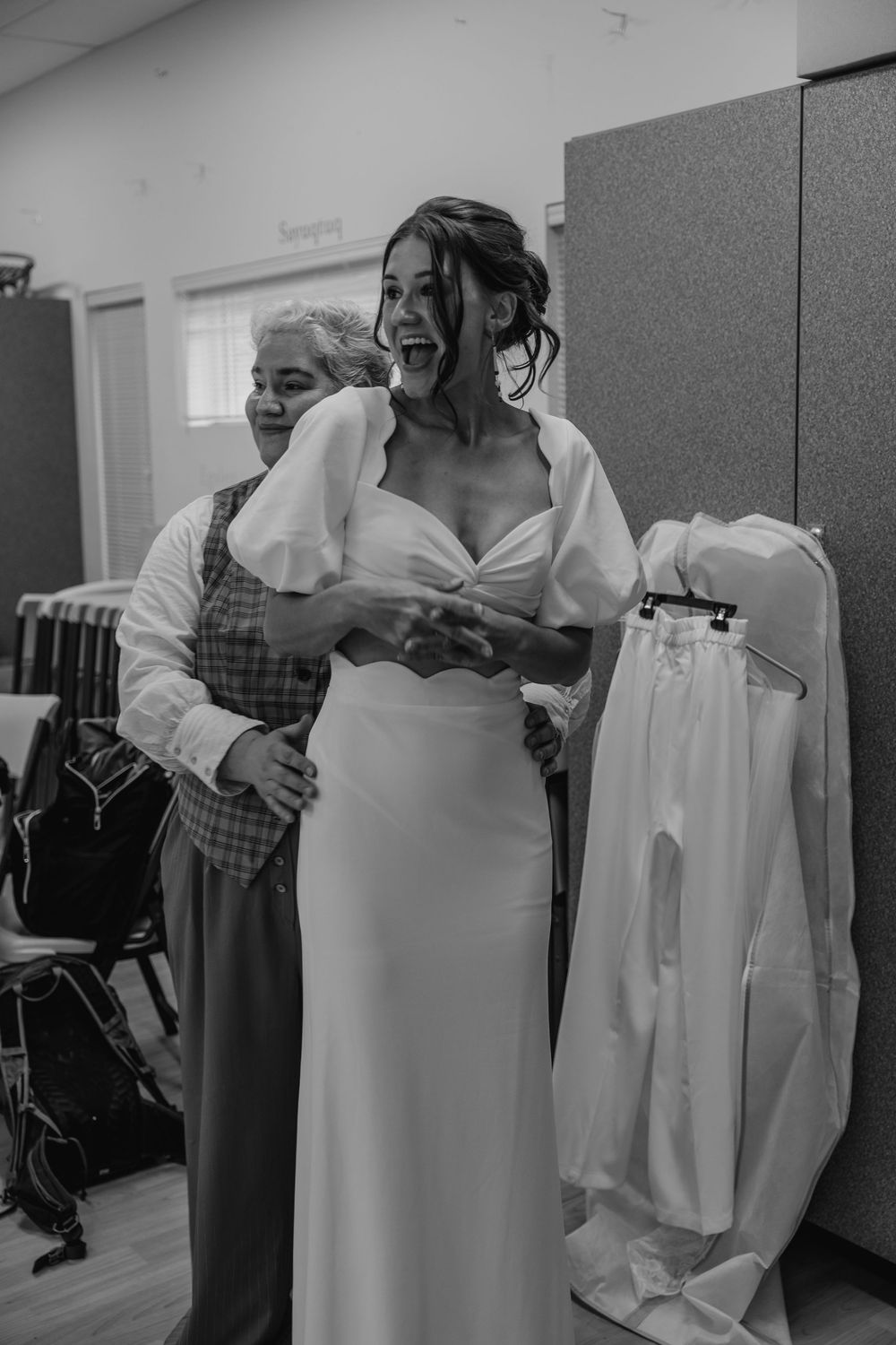 Black and white photo of a joyful bride in a white wedding dress being embraced by an older woman.