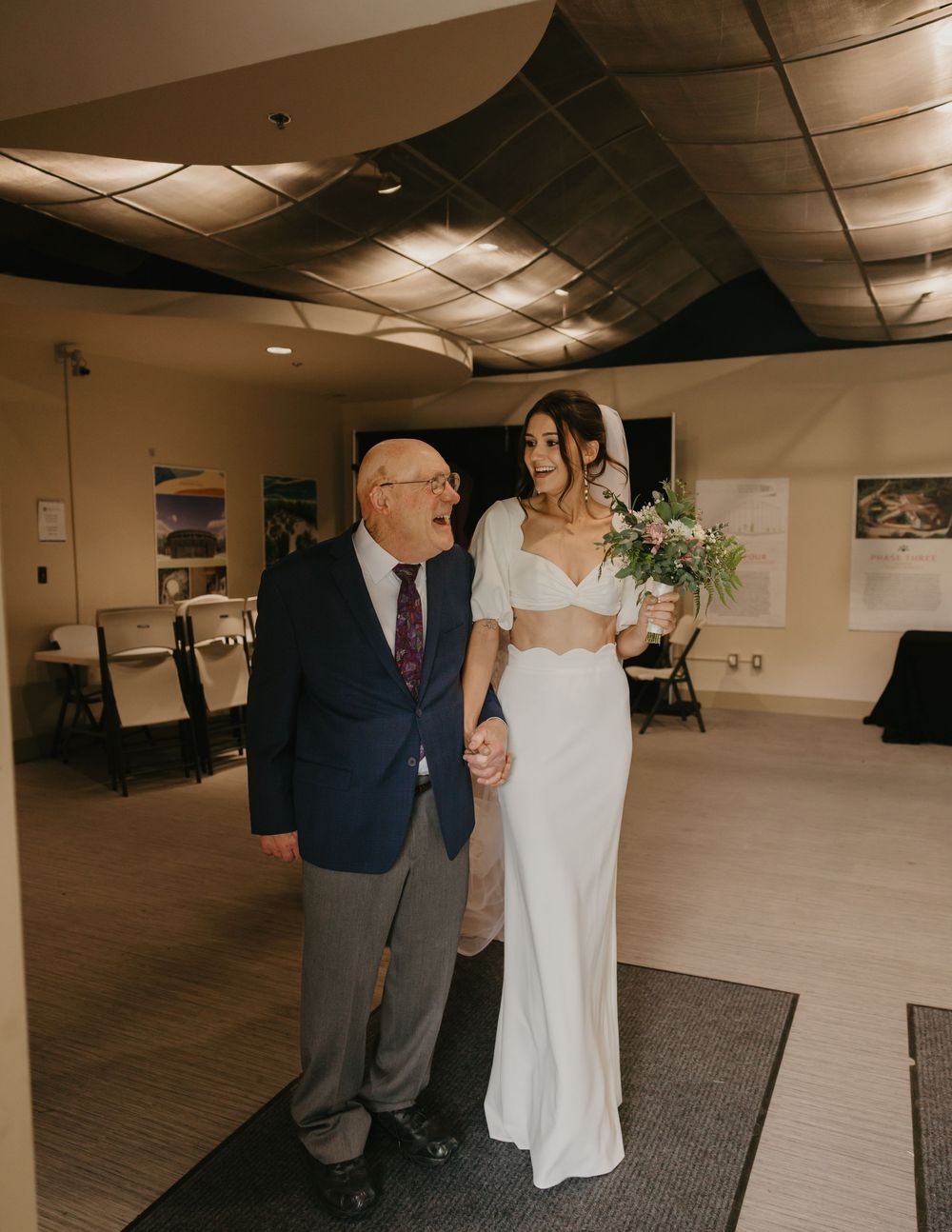 Bride in two-piece white wedding gown holding bouquet while walking with elderly grandfather indoors.