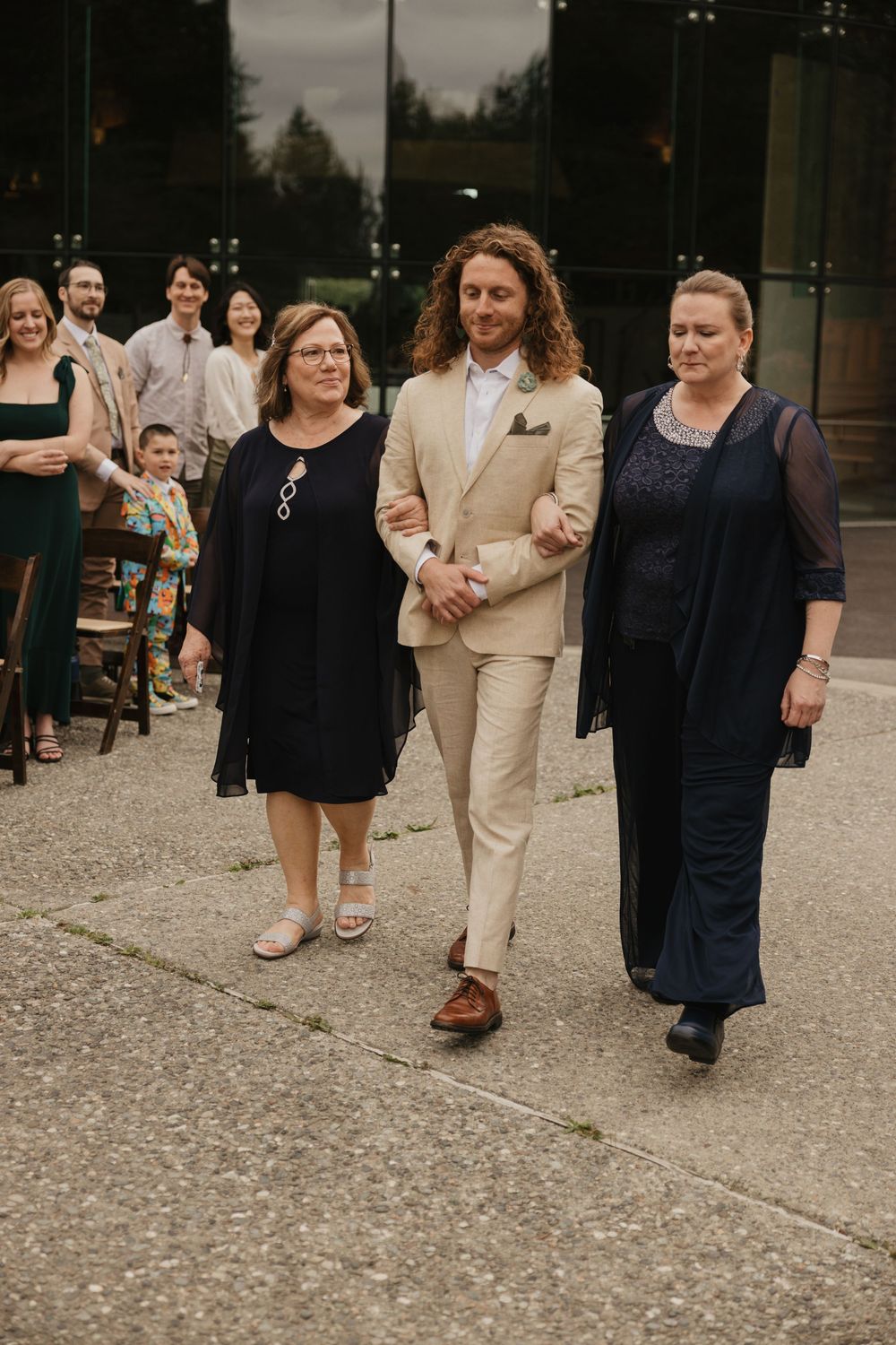 Groom in beige suit being escorted down the aisle by two women at an outdoor wedding ceremony.