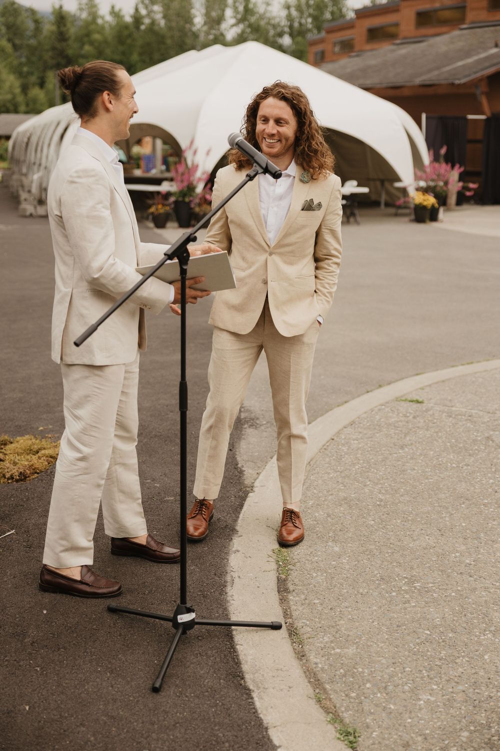 Two men in matching beige suits sharing a joyful moment at an outdoor wedding ceremony.