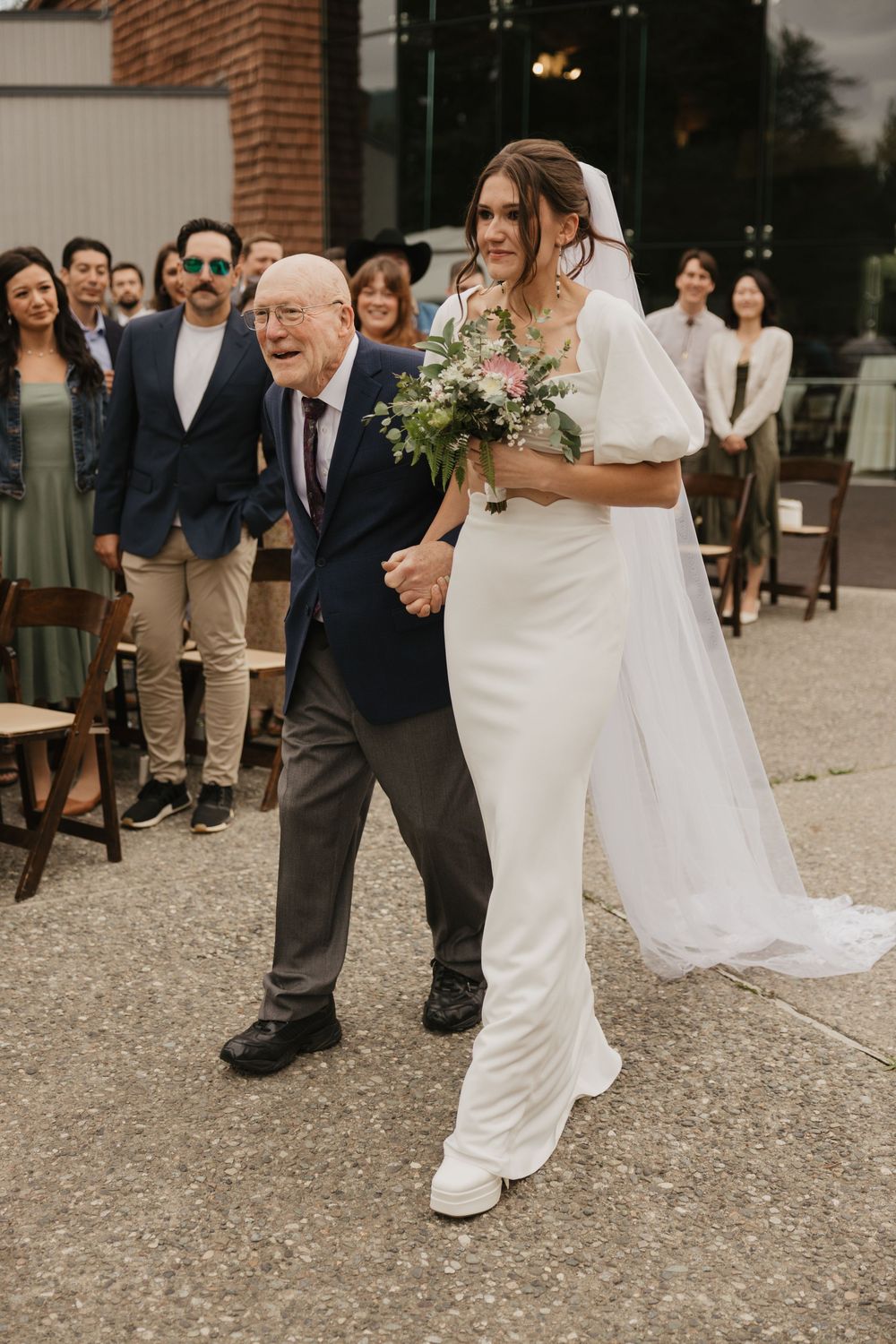Radiant bride in white gown and veil walking down the aisle with her grandfather at outdoor wedding.