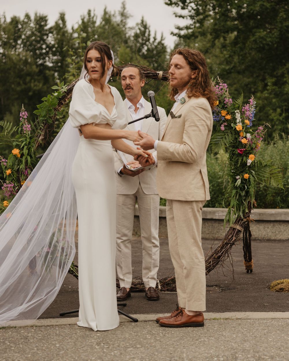 Bride and groom holding hands during outdoor wedding ceremony with floral arch and officiant behind them.