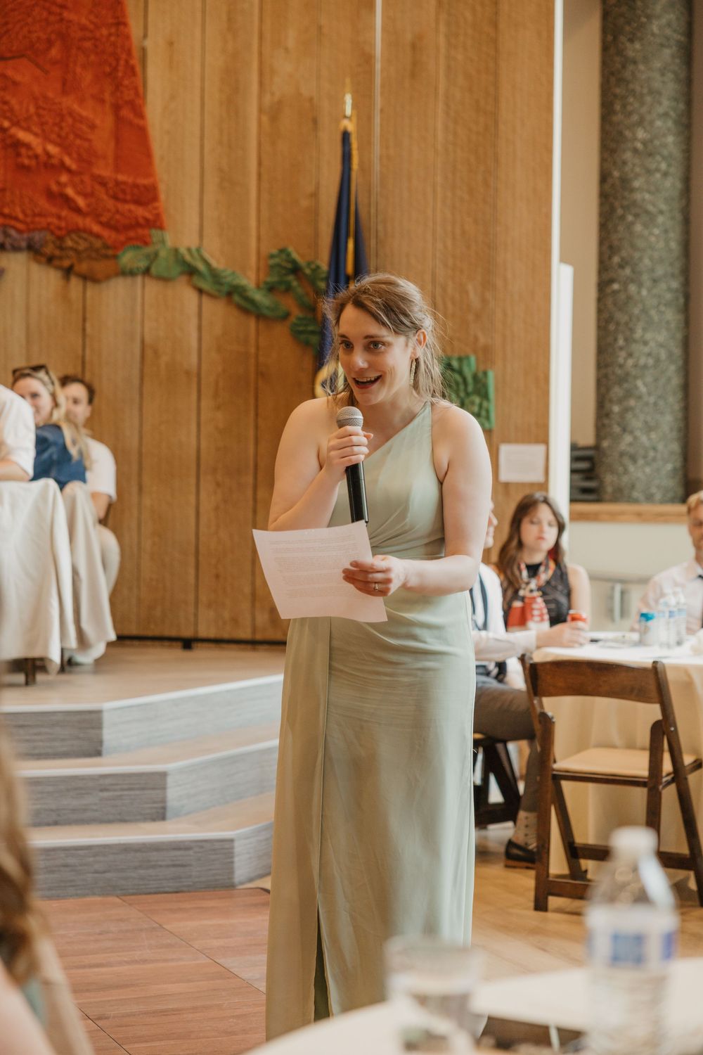 Bridesmaid in sage green gown delivering a heartfelt speech with microphone at wedding reception.
