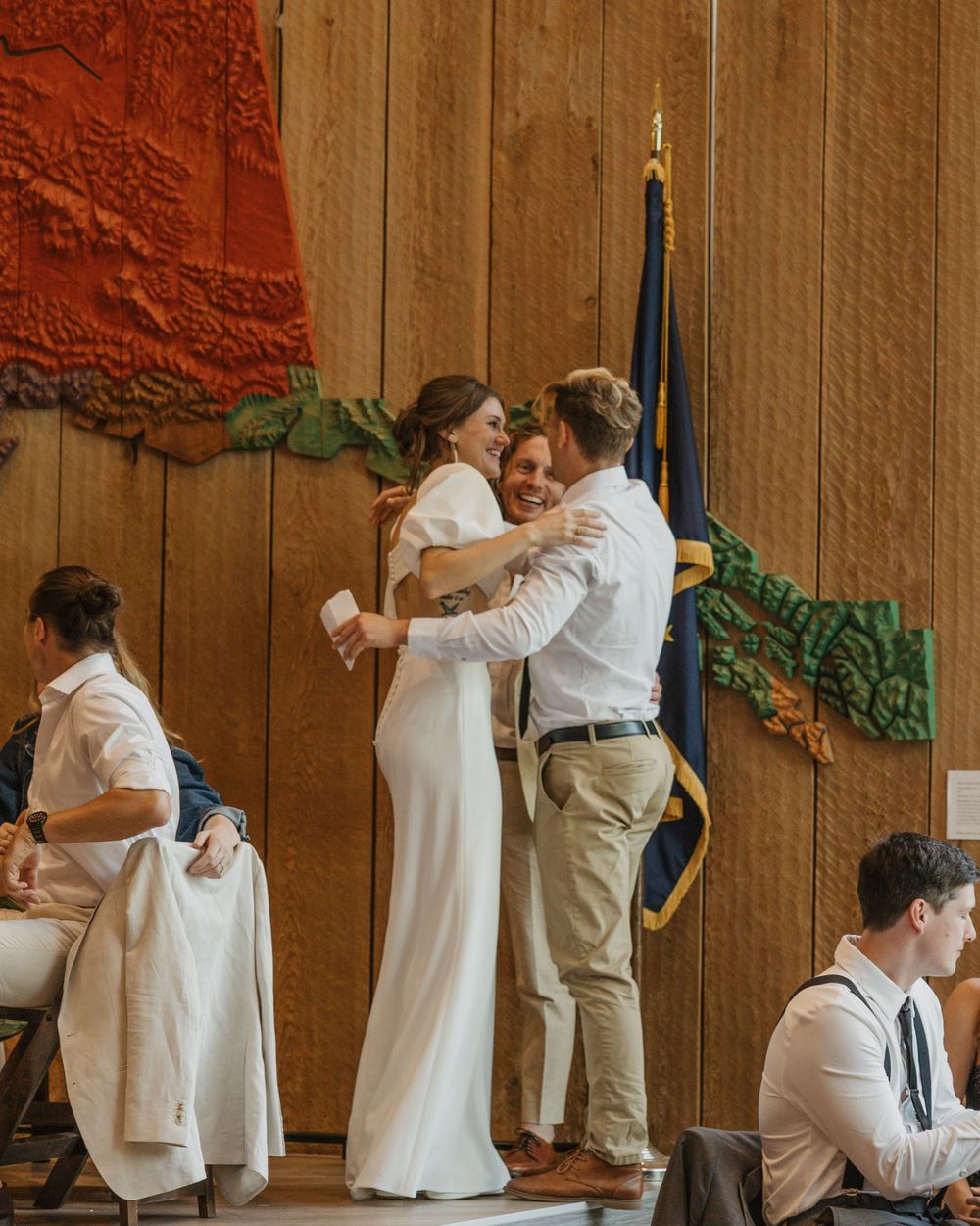 Bride in white gown sharing a joyful hug with a guest at a rustic wood-paneled wedding reception.