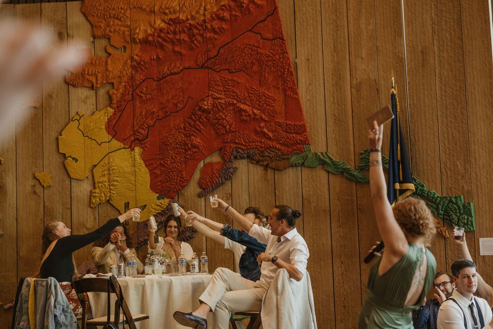 Wedding guests raising glasses in a toast at reception with colorful Alaska map mural backdrop.