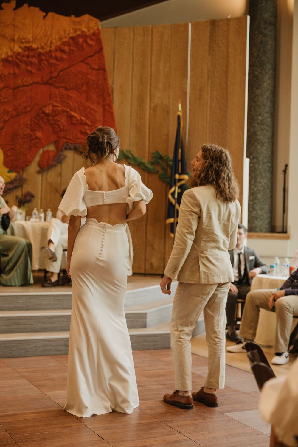 Bride in white gown with button-back detail stands beside groom in beige suit at wedding reception.