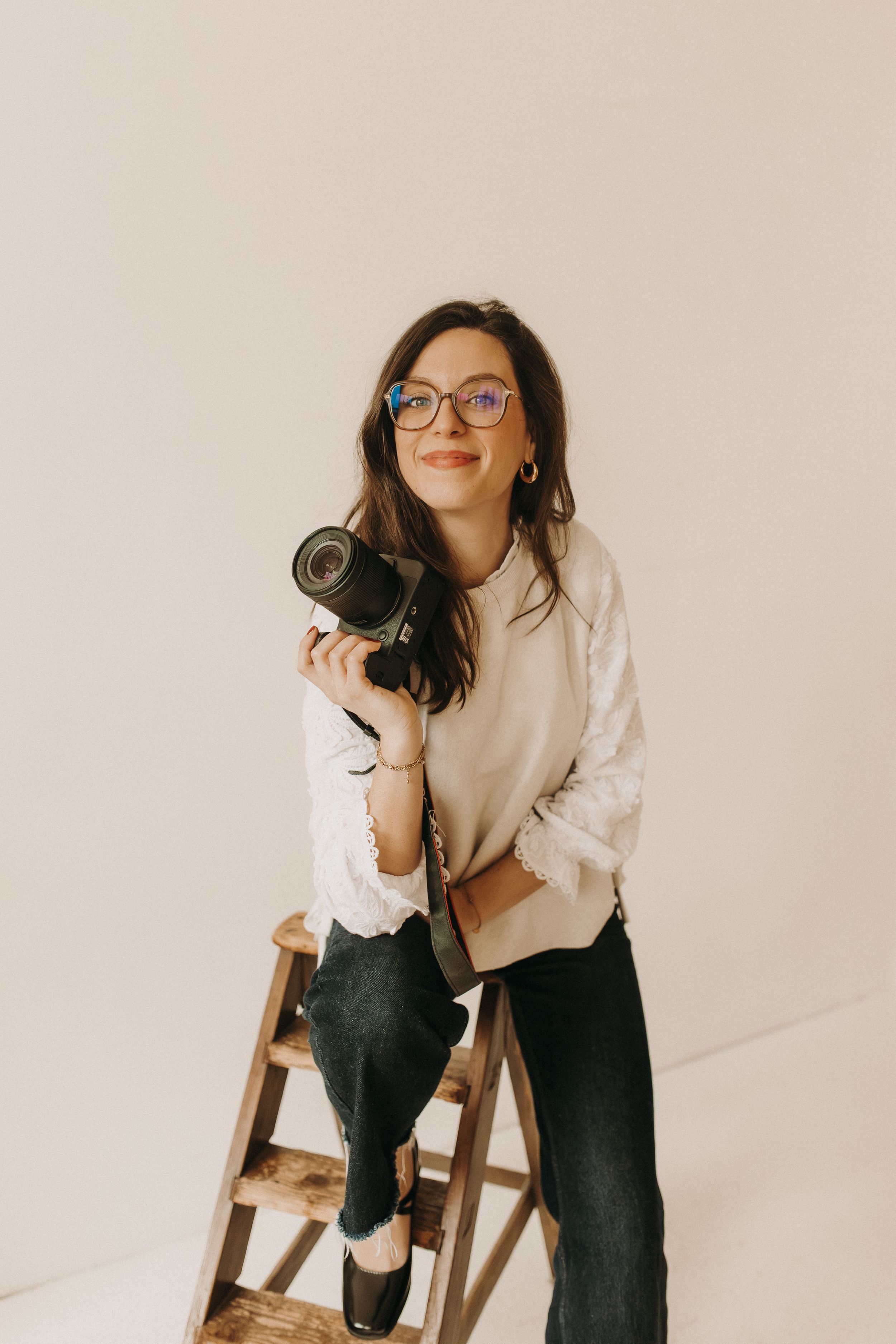 A person in glasses and a white sweater poses casually on a wooden chair against a light background.