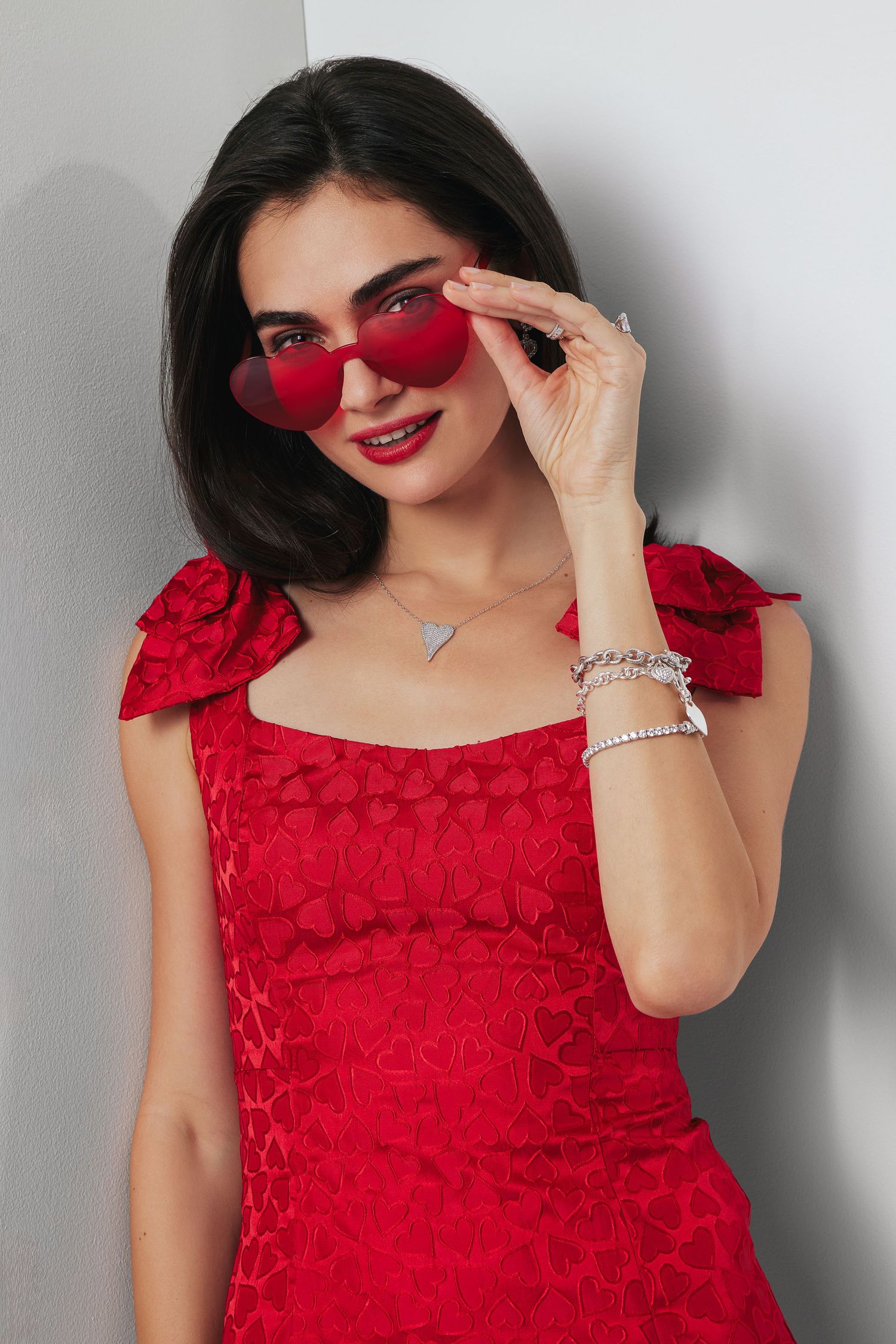 Studio fashion portrait featuring a woman in a red dress with heart details, red sunglasses, and silver jewelry on a light background.