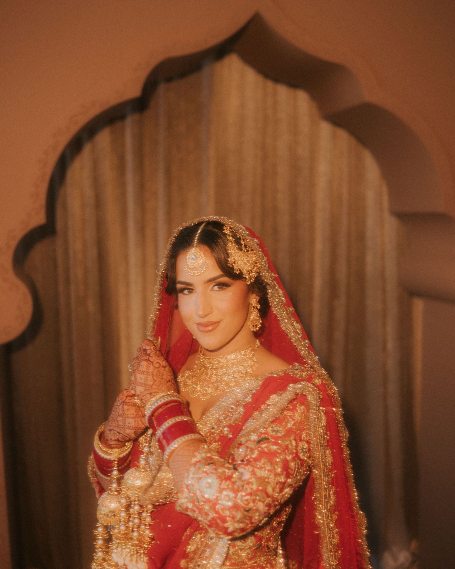A bride in traditional South Asian wedding attire wearing a red and gold embroidered lehenga with ornate jewelry.
