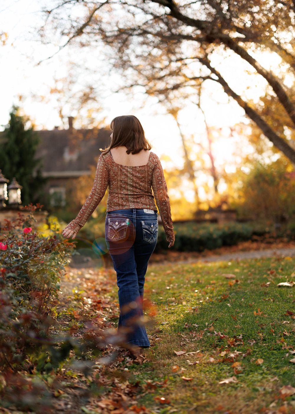 Person walking on autumn path wearing brown sweater and jeans against golden sunset backdrop.