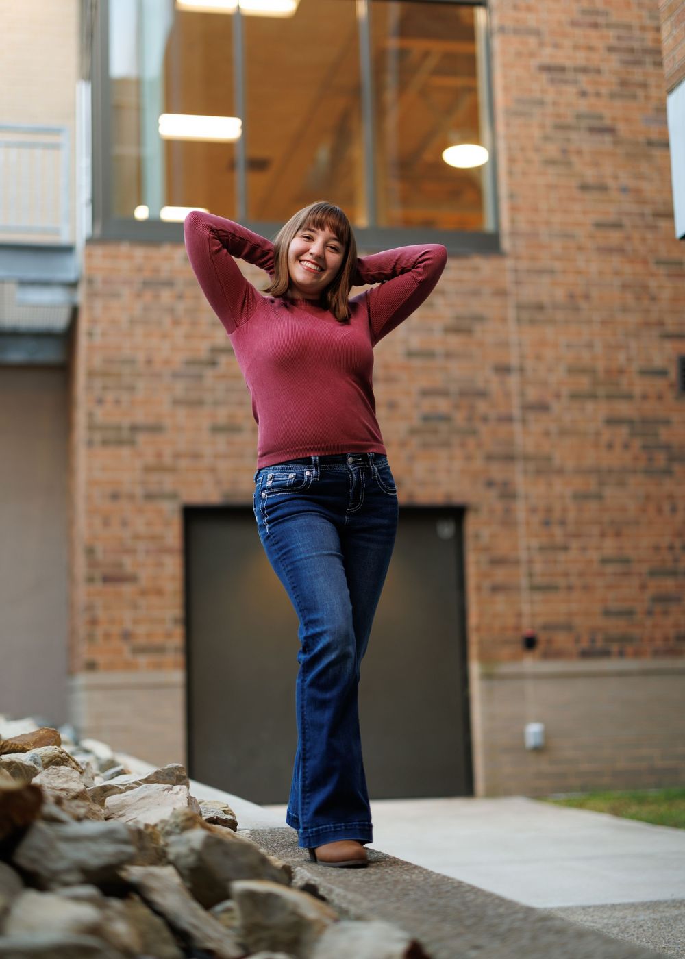 Person in burgundy sweater and jeans posing playfully by brick building.