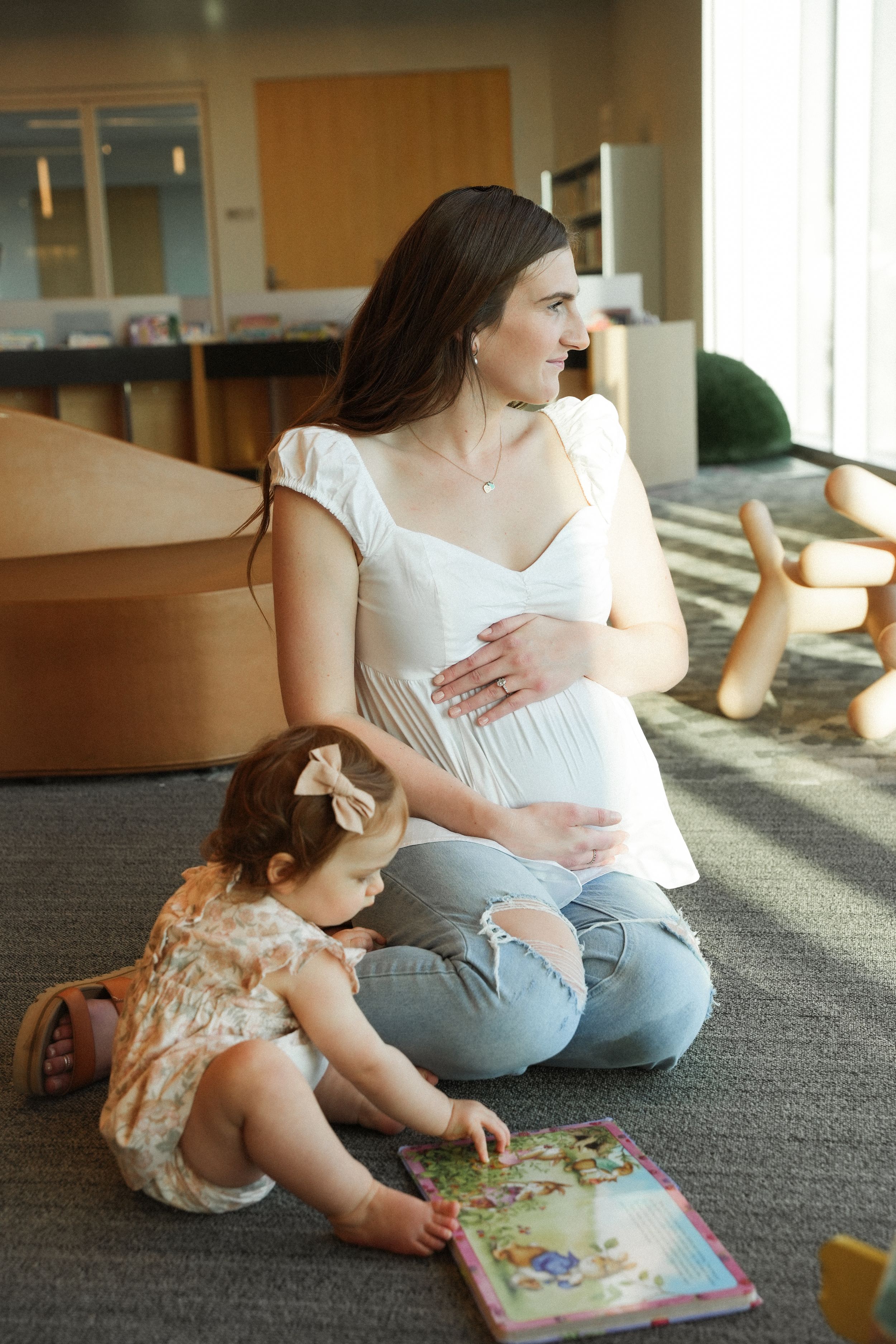A parent and toddler sit on carpeted floor looking at a colorful children's book together in sunlit room.
