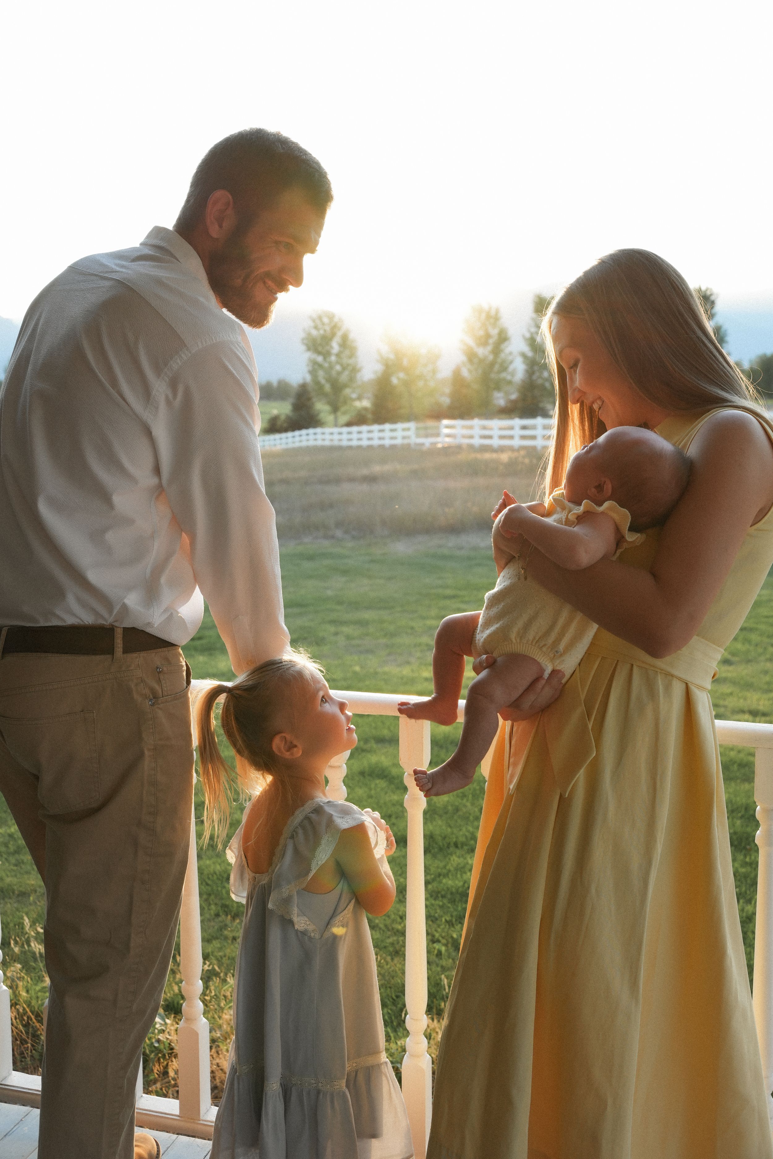 Family sharing tender moment together on porch at sunset with white fence and trees in background.