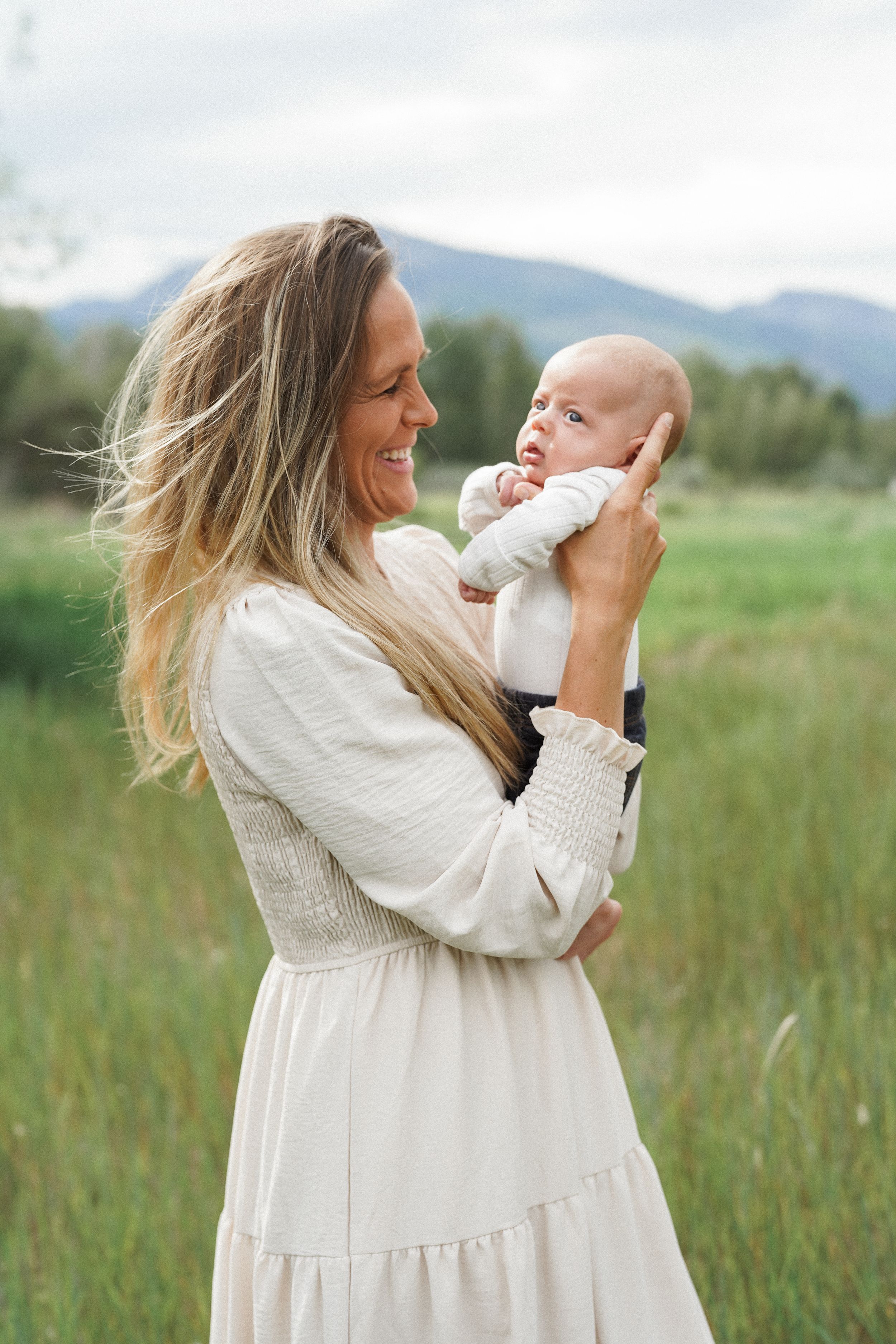 A mother in a white dress holds and plays with her baby in a grassy field with mountains in the background.