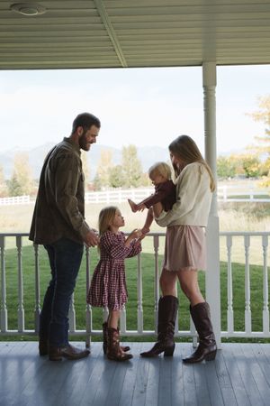 Family of four sharing a warm moment on a farmhouse porch, with white fence and mountain views in background.