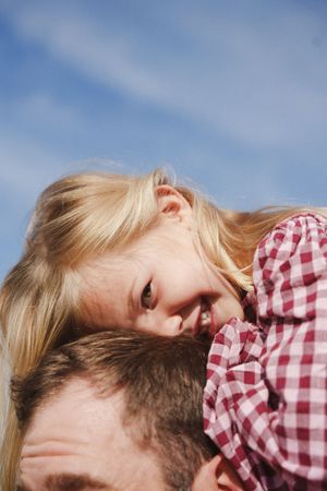 Young blonde girl in red plaid shirt resting her head on a man's shoulder against a blue sky.