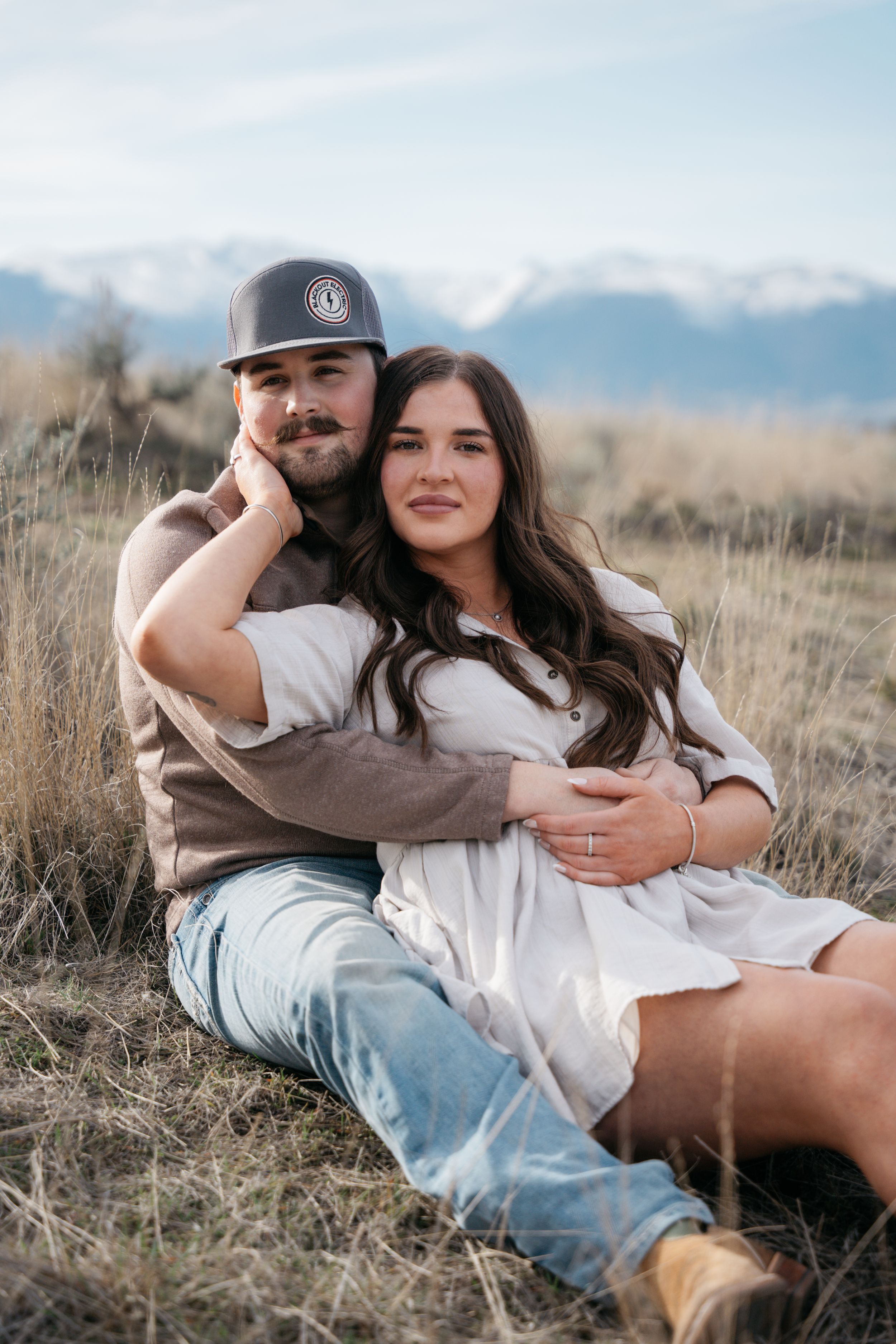 A couple sits together in a grassy field with snow-capped mountains, the man wearing a cap embracing a woman in a white dress.