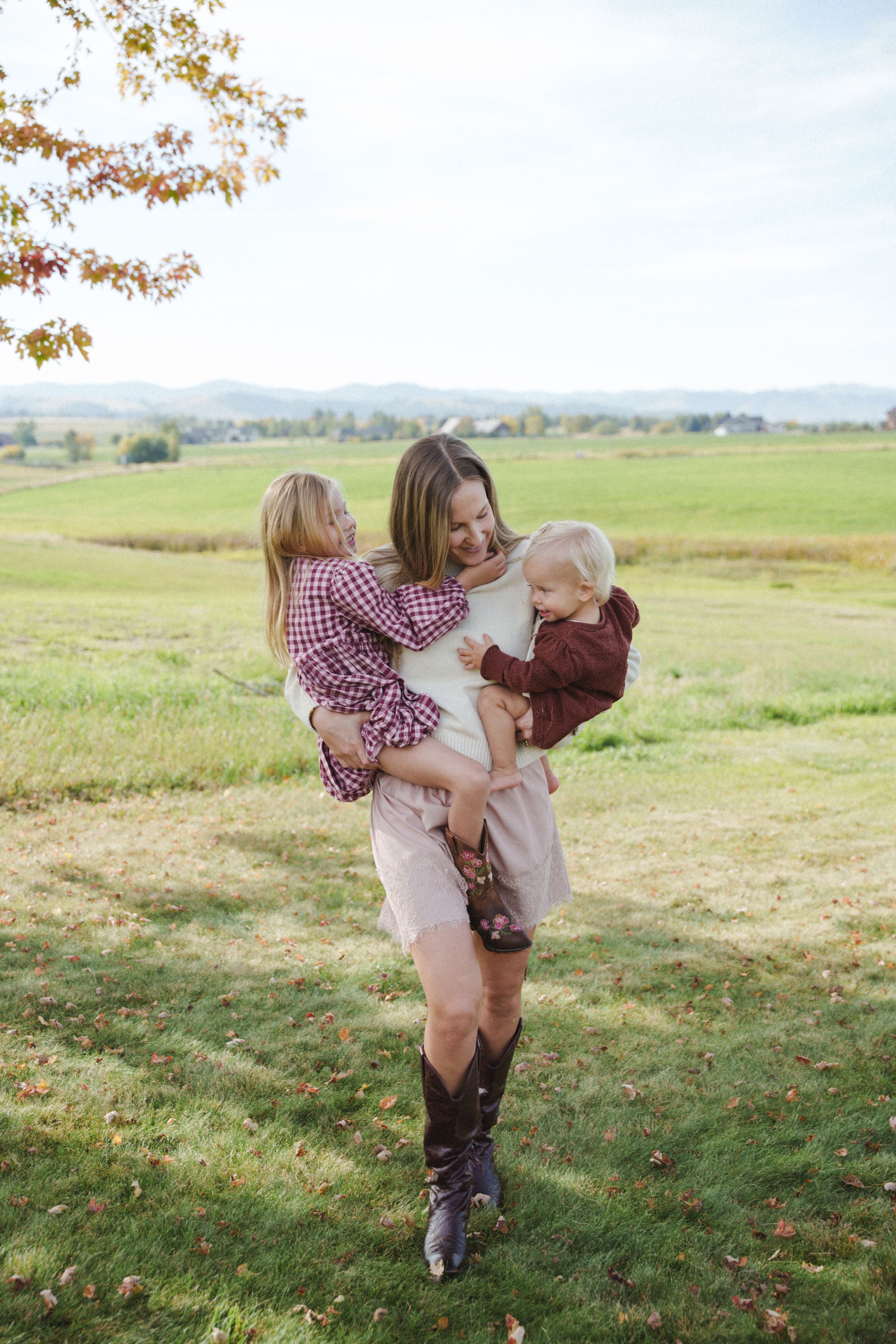 A smiling mother holds two young children in a grassy field, wearing cowboy boots and casual clothes on a sunny day.