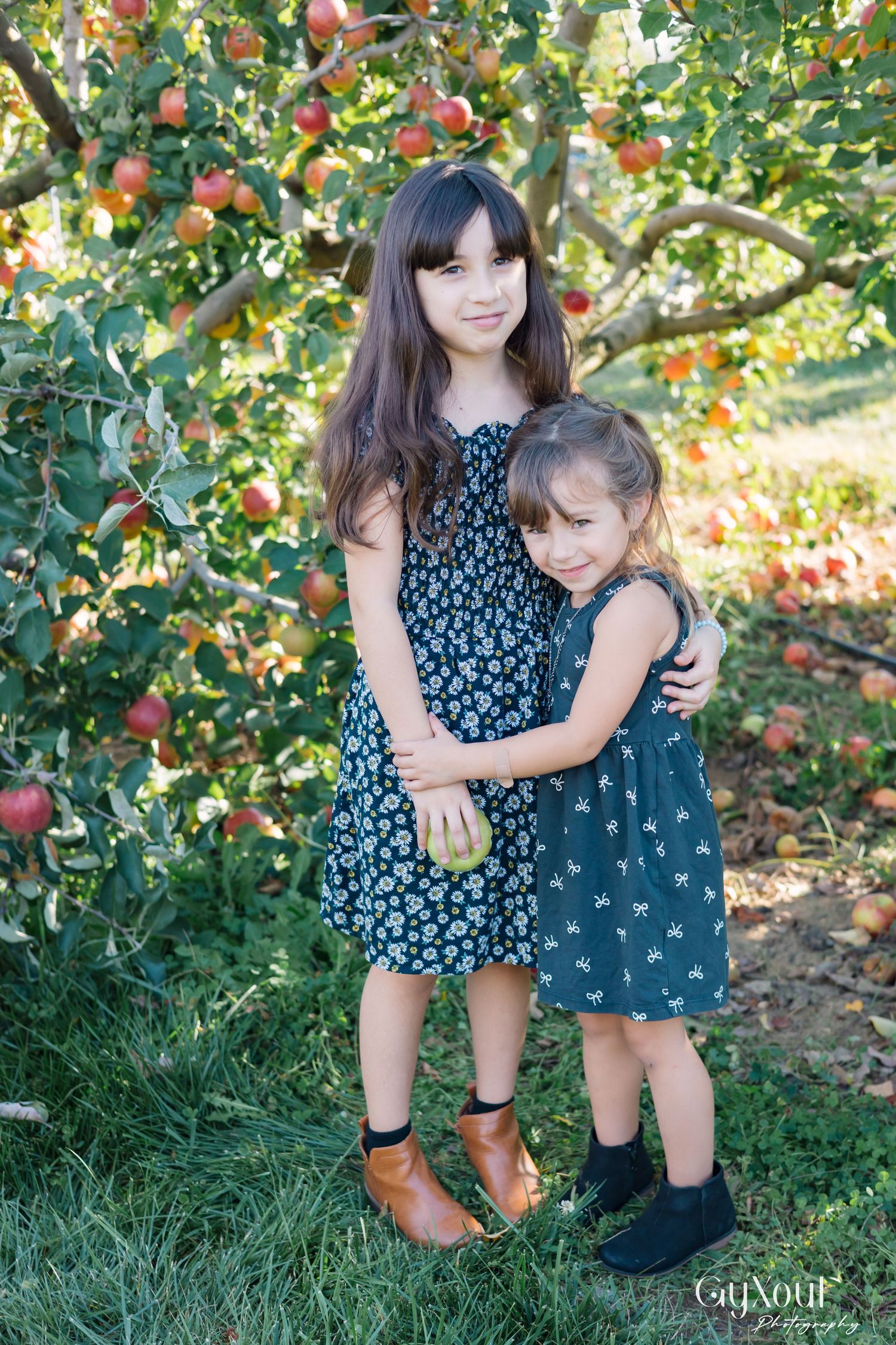 Two children in floral dresses play together in an apple orchard on a sunny autumn day.