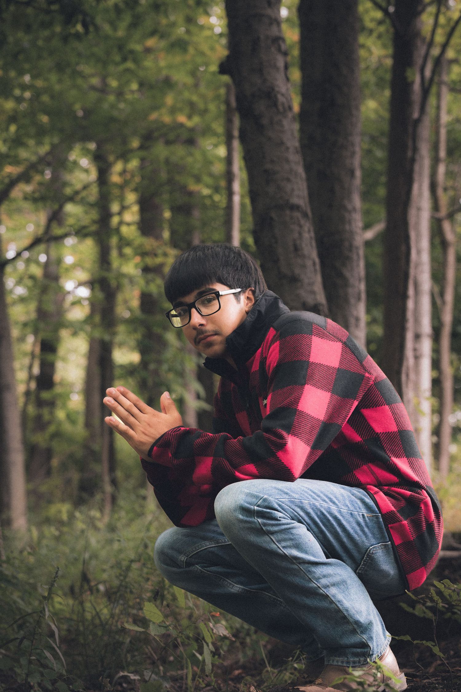 Person in red plaid shirt and jeans crouching in autumn forest setting.
