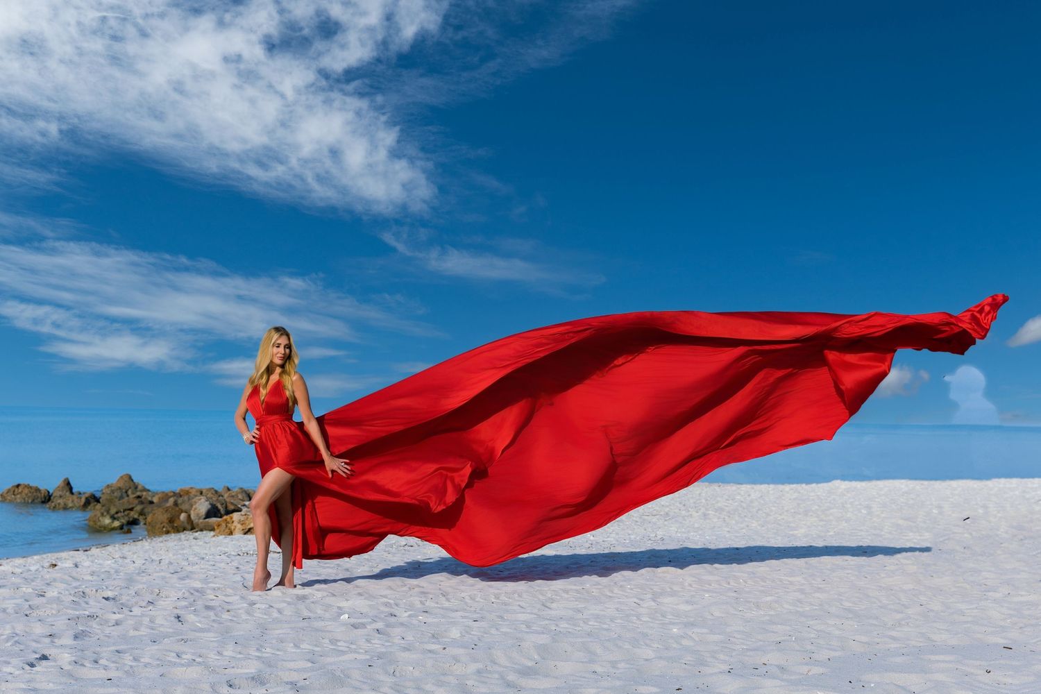 Figure in flowing red dress stands on white sand with billowing fabric against bright blue sky and clouds.